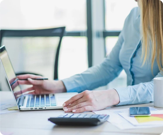 Mulher sentada usando um notebook, na mesa possui calculadora, papéis e celular.